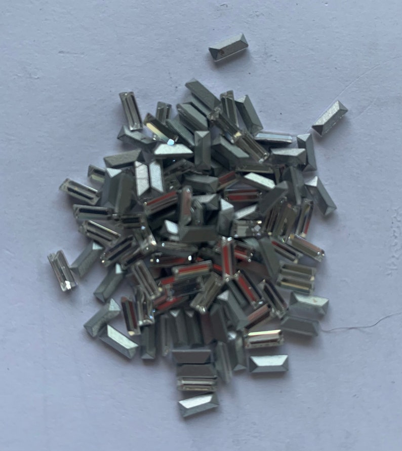 May include: A pile of silver-colored, rectangular-shaped rhinestones. The rhinestones are scattered on a white surface.