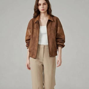 Peut inclure: Une femme porte une veste en daim marron avec un col et une fermeture éclair sur le devant. Elle porte également un haut blanc et un pantalon beige. La tenue est complétée par des baskets blanches.