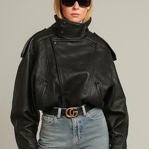 May include: A black leather jacket with a high collar and asymmetrical zipper design. The jacket features shoulder epaulets and a wide belt with a gold buckle. The model is wearing sunglasses and blue jeans. This is a stylish outerwear piece.