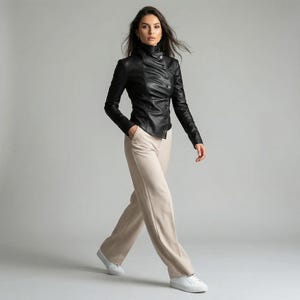May include: A black leather jacket with a high collar and button closure. The jacket is paired with beige pants and white sneakers. The outfit is a modern, stylish ensemble.