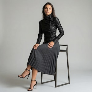 May include: A woman wearing a black leather jacket with a high collar and a gray pleated skirt. She is seated on a dark metal stool, wearing black high-heeled sandals. The outfit is a blend of classic and modern styles.