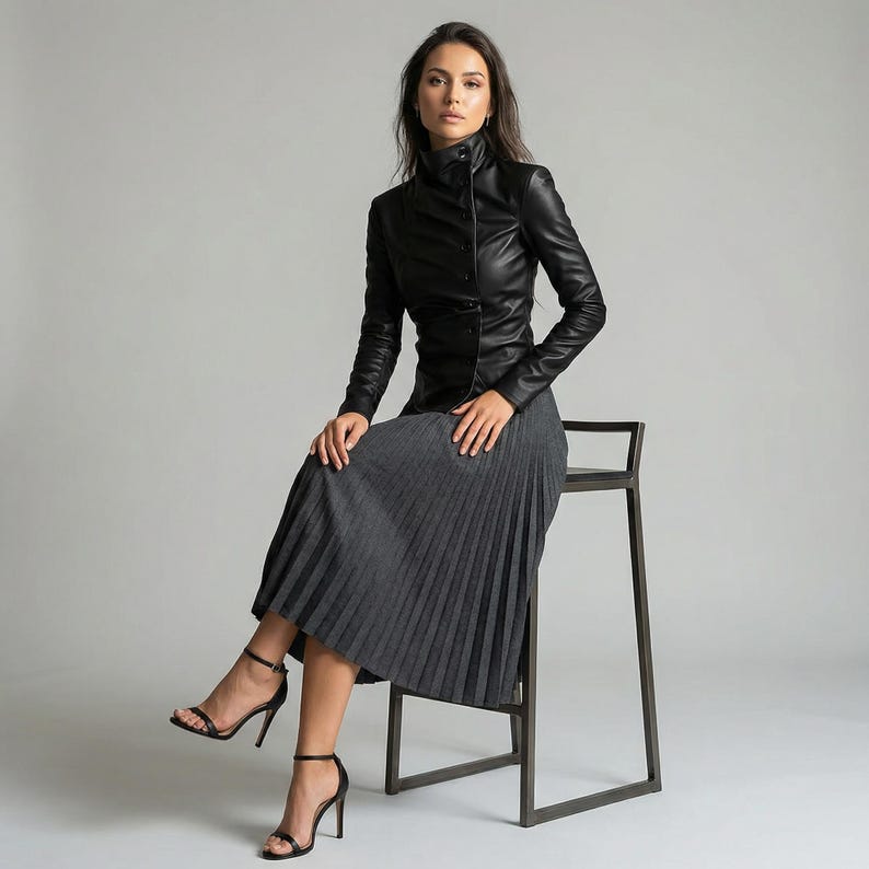 May include: A woman wearing a black leather jacket with a high collar and a gray pleated skirt. She is seated on a dark metal stool, wearing black high-heeled sandals. The outfit is a blend of classic and modern styles.