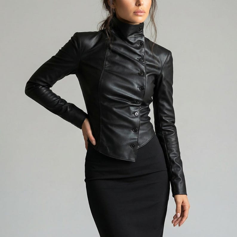 May include: A black leather jacket with a high collar and button closure. The jacket has long sleeves and a fitted silhouette. The model is wearing a black skirt. The jacket is a stylish outerwear piece.