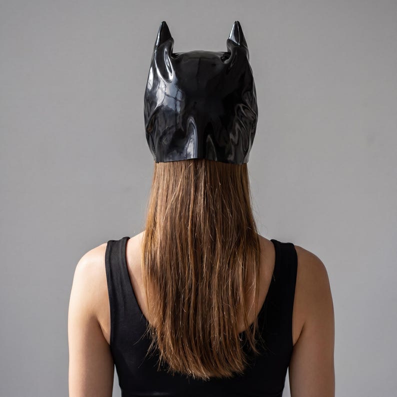 May include: A black, glossy mask with pointed ears, resembling a bat, is worn by a person. The mask covers the top of the head and forehead. The person is wearing a black tank top. The background is a plain, light gray.