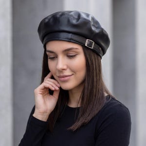 May include: A black leather beret with a silver buckle detail. The beret is round with a slightly raised crown and a smooth, shiny texture. The hat is a classic style, suitable for various outfits and occasions.