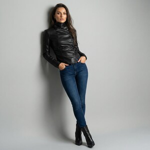 May include: A black leather jacket with a high collar and button details. The jacket is paired with dark blue jeans and black ankle boots. The model is leaning against a gray wall.