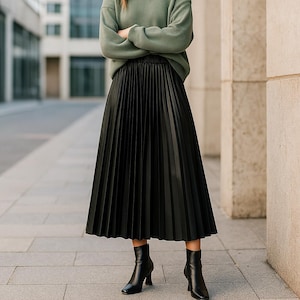 May include: A black, pleated midi skirt with a high waist. The skirt is paired with black heeled ankle boots. The person is wearing a green sweater with their arms crossed.