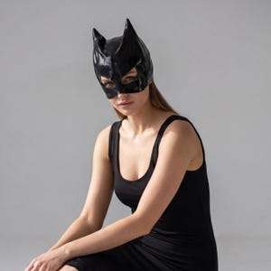 May include: A black cat-eared mask with a glossy finish. The mask covers the eyes and forehead, featuring pointed ears. The person in the image is wearing a black tank top dress. The background is a plain, light gray.