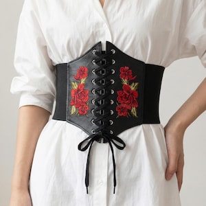 Puede incluir: Cinturón corsé negro con bordado floral rojo. El corsé presenta un frente con cordones y una banda elástica ancha. Las flores bordadas añaden un toque decorativo a este accesorio que ciñe la cintura.