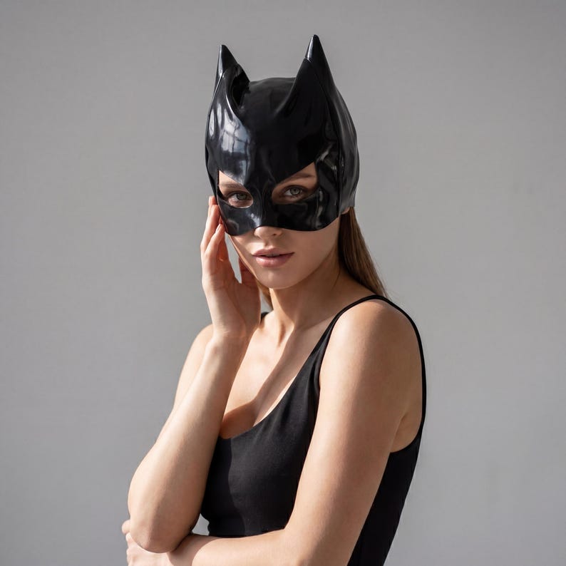 May include: A black cat-eared mask with eye cutouts. The mask is made of a shiny material and covers the upper face. The person in the image is wearing a black tank top.