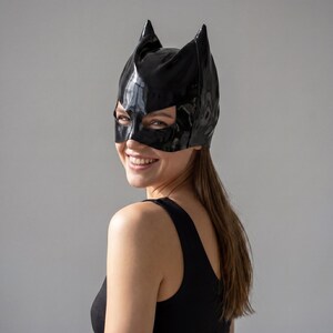 May include: A glossy black cat-eared mask. The mask covers the eyes and nose, with pointed ears extending upwards. The mask is worn on the head. The overall theme is costume and cosplay.