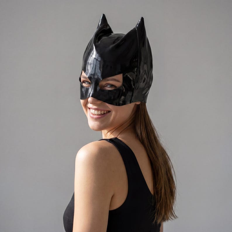 May include: A glossy black cat-eared mask. The mask covers the eyes and nose, with pointed ears extending upwards. The mask is worn on the head. The overall theme is costume and cosplay.