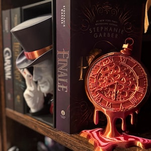 May include: A decorative bookshelf with a top hat bookend, a book titled "FINALE" by Stephanie Garber, and a red and gold clock-shaped ornament. The clock ornament has a dripping design and Roman numerals.