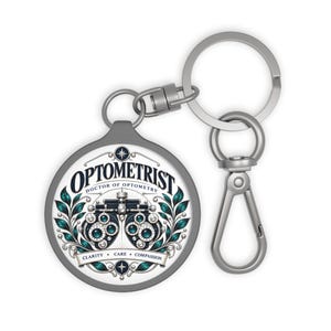May include: A round silver keychain featuring a graphic of a phoropter, the text OPTOMETRIST DOCTOR OF OPTOMETRY, and the words CLARITY, CARE, COMPASSION. The design includes teal and black leaf accents and a silver metal lobster clasp.