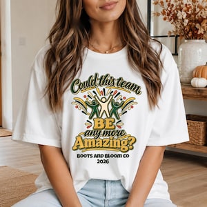 Puede incluir: Una camiseta blanca de manga corta con un gráfico que incluye el texto "Could this team BE any more Amazing?" en verde y amarillo, acompañado de tres figuras estilizadas y el texto "BOOTS AND BLOOM CO 2026" impreso en negro en la parte inferior.