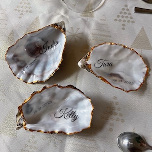 May include: Three oyster shells with gold rims, each inscribed with a name and date in black script: Indra, Tara, and Kelly, with the date 28-11-2025. They are arranged on a white tablecloth. A spoon, fork, and wine glass are also visible.