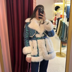 Puede incluir: Un abrigo de lentejuelas azul y verde con ribete de piel sintética blanca y cinturón a juego. La persona lleva vaqueros azul oscuro y mocasines negros. El abrigo es el punto focal de la imagen.