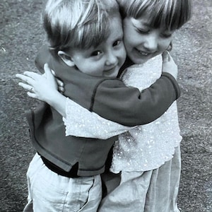 May include: Black and white photograph of two young children embracing. One child wears a dark red long-sleeved shirt and light-colored pants. The other child wears a light-colored top and skirt. Both children are smiling.