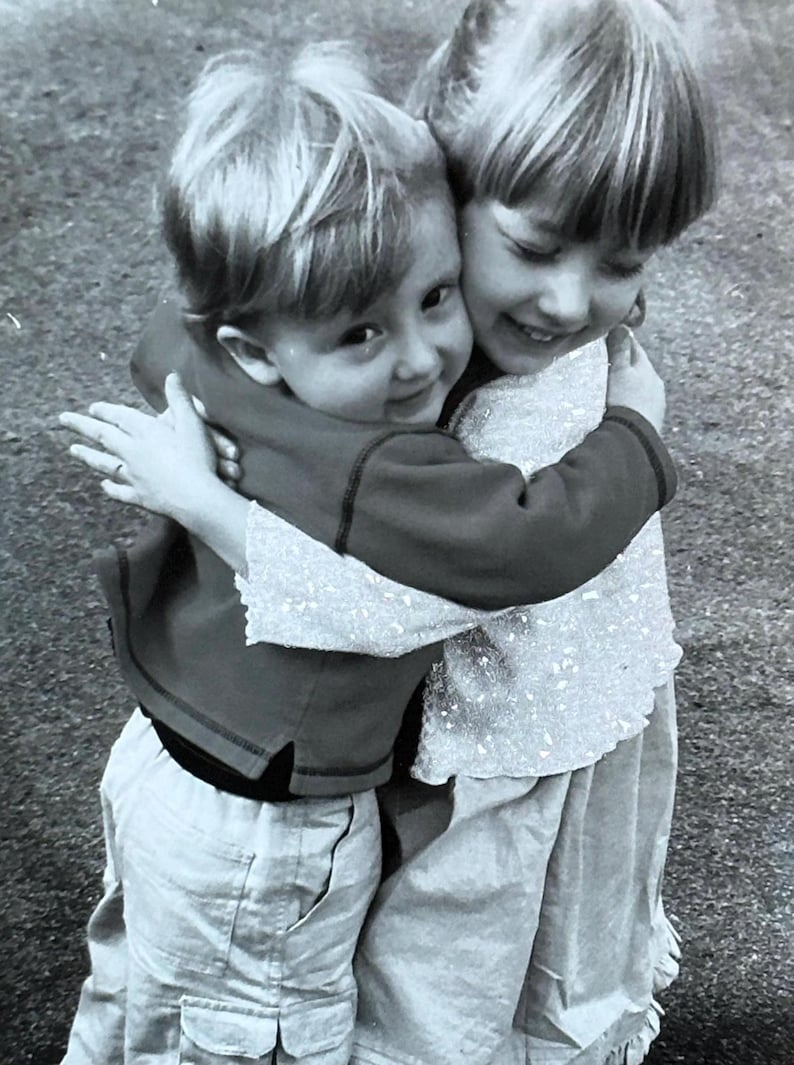 May include: Black and white photograph of two young children embracing. One child wears a dark red long-sleeved shirt and light-colored pants. The other child wears a light-colored top and skirt. Both children are smiling.