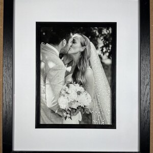 May include: Black and white framed photograph of a couple kissing, with the bride holding a bouquet of white flowers. The photograph is set within a white mat and a black wooden frame.