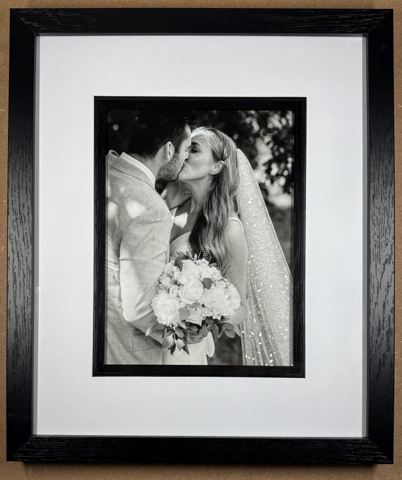 May include: Black and white framed photograph of a couple kissing, with the bride holding a bouquet of white flowers. The photograph is set within a white mat and a black wooden frame.