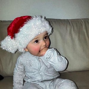 May include: A baby wearing a red and white Santa hat and a gray onesie with a pattern of small, light gray images. The baby is sitting on a beige couch and has rosy cheeks.