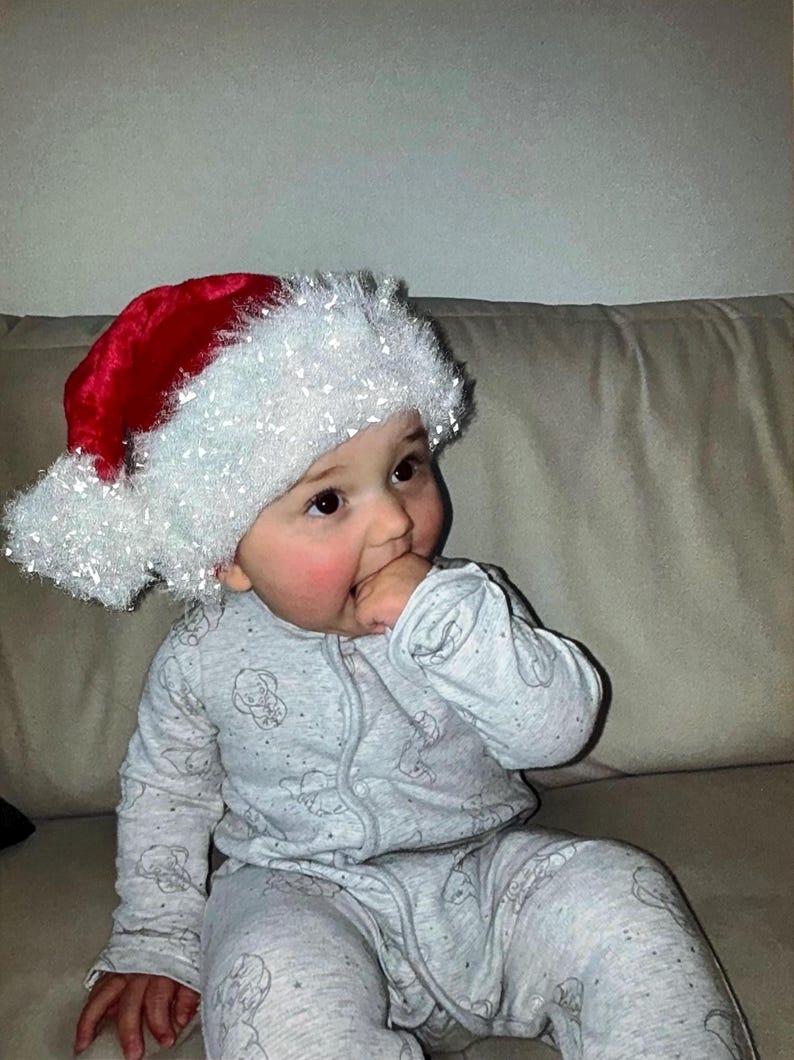 May include: A baby wearing a red and white Santa hat and a gray onesie with a pattern of small, light gray images. The baby is sitting on a beige couch and has rosy cheeks.
