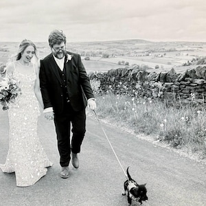 May include: Black and white photo of a newly married couple walking down a road, holding hands, with a small dog on a leash. The bride wears a beaded gown and carries a bouquet. The groom is in a suit. A stone wall and rolling hills are in the background.