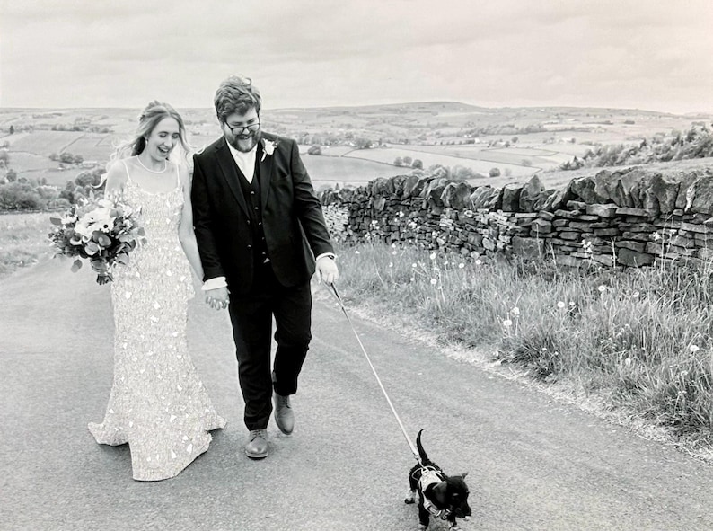 May include: Black and white photo of a newly married couple walking down a road, holding hands, with a small dog on a leash. The bride wears a beaded gown and carries a bouquet. The groom is in a suit. A stone wall and rolling hills are in the background.