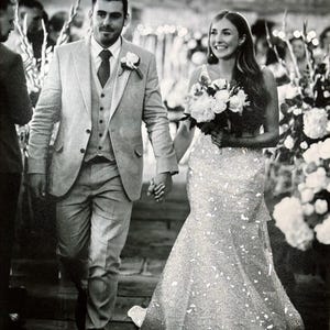 May include: Black and white photo of a wedding. A person in a light gray suit and a person in a sparkling white wedding dress walk hand-in-hand. The person in the dress carries a bouquet of white flowers.