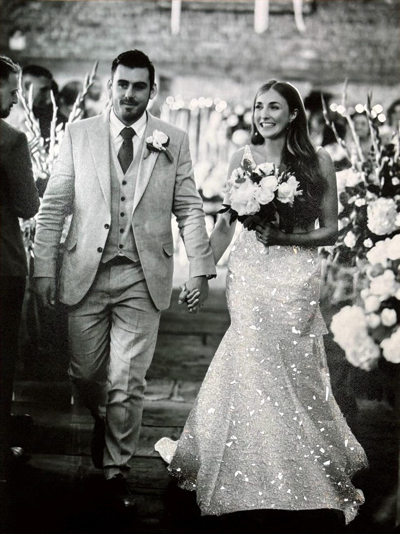 May include: Black and white photo of a wedding. A person in a light gray suit and a person in a sparkling white wedding dress walk hand-in-hand. The person in the dress carries a bouquet of white flowers.