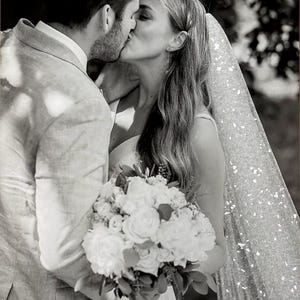 May include: Black and white photo of a couple kissing. The woman wears a veil and holds a bouquet of white flowers. The man wears a light-colored suit. The image captures a moment of intimacy and celebration, likely at a wedding.