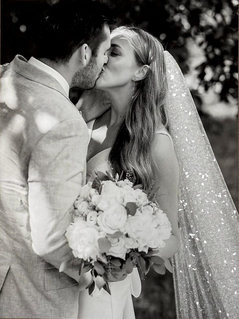 May include: Black and white photo of a couple kissing. The woman wears a veil and holds a bouquet of white flowers. The man wears a light-colored suit. The image captures a moment of intimacy and celebration, likely at a wedding.