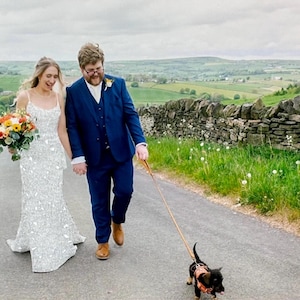 May include: A newly married couple walks down a road, holding hands, with a small dog on a leash. The bride wears a white, beaded wedding dress and carries a bouquet. The groom wears a blue suit. The scene is set in a rural landscape.