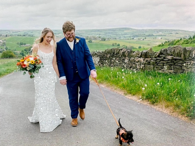 May include: A newly married couple walks down a road, holding hands, with a small dog on a leash. The bride wears a white, beaded wedding dress and carries a bouquet. The groom wears a blue suit. The scene is set in a rural landscape.