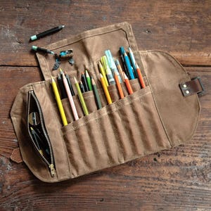 May include: A brown canvas pencil roll with a variety of colored pencils, pens, and a pencil sharpener. The roll is open, displaying the contents in individual pockets. A leather strap secures the roll when closed. The background is a wooden surface.