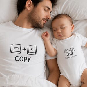 Puede incluir: Una camiseta blanca con "ctrl + c COPY" impresa en negro, junto a un bebé con un body blanco con "ctrl + v PASTE (improving version)" impreso en negro. Ambos están acostados en una cama blanca.