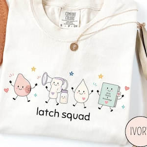 May include: An ivory t-shirt featuring a graphic of four cartoon characters: a breast, a breast pump with a bottle, a milk drop, and a book titled Milk Moms & Knowledge. Below the characters, the text reads latch squad in black lowercase letters.
