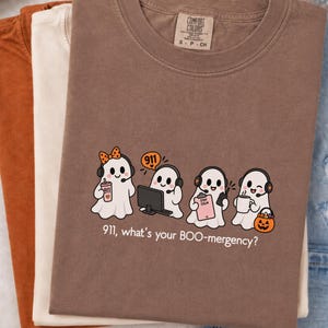 Peut inclure: Un t-shirt marron avec un motif de quatre fantômes de dessin animé portant des casques. Les fantômes sont représentés comme des répartiteurs d'urgence avec un ordinateur, un presse-papiers et un seau citrouille. Le texte indique, 911, what's your BOO-mergency?