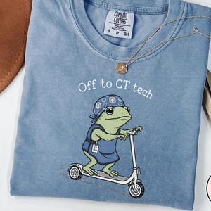 May include: A blue jean colored t-shirt featuring a graphic of a green frog wearing a blue bandana and a blue scrub top, riding a white scooter. The text above the frog reads Off to CT tech. The shirt has a Comfort Colors brand label at the neck.