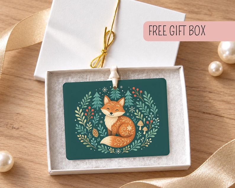 May include: A rectangular ornament featuring a fox illustration on a dark green background, surrounded by a wreath of pine trees, berries, and leaves. The ornament is presented inside a white gift box with a gold ribbon and a FREE GIFT BOX label.