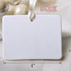 May include: A rectangular white ceramic ornament with rounded corners and a hole at the top for hanging. A cream-coloured ribbon is threaded through the hole. The ornament measures 3 inches in height, 4 inches in width, and 4 mm in thickness.