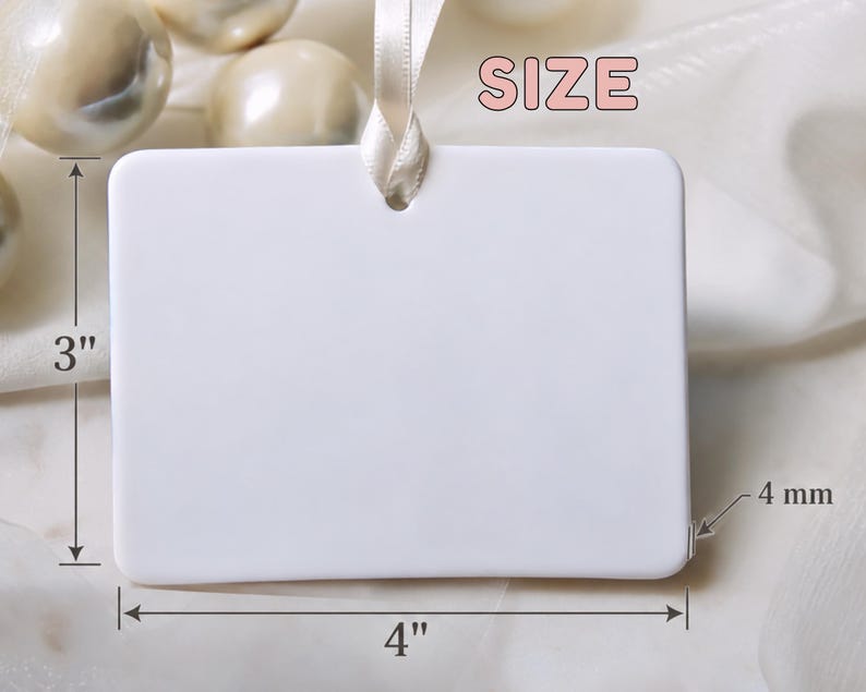 May include: A rectangular white ceramic ornament with rounded corners and a hole at the top for hanging. A cream-coloured ribbon is threaded through the hole. The ornament measures 3 inches in height, 4 inches in width, and 4 mm in thickness.