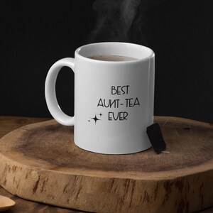 May include: A white ceramic mug with the text "BEST AUNT-TEA EVER" in black. Steam rises from the mug, suggesting a hot beverage. A tea bag is attached to the side. The mug sits on a wooden surface.
