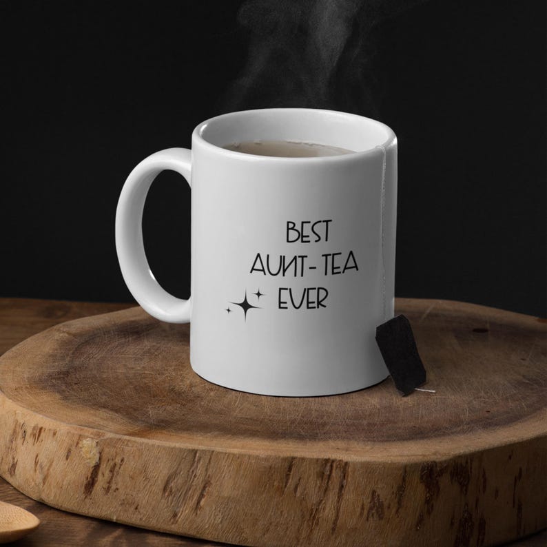 May include: A white ceramic mug with the text "BEST AUNT-TEA EVER" in black. Steam rises from the mug, suggesting a hot beverage. A tea bag is attached to the side. The mug sits on a wooden surface.