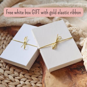 May include: Two rectangular white gift boxes, each secured with a thin gold elastic ribbon tied in a small bow. The text Free white box GIFT with gold elastic ribbon is displayed above the boxes, which are positioned on a textured surface.