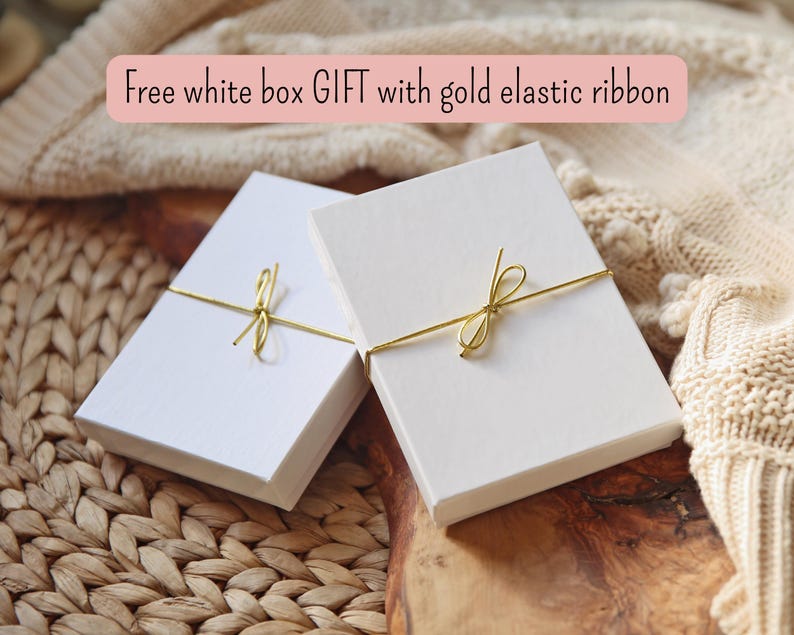 May include: Two rectangular white gift boxes, each secured with a thin gold elastic ribbon tied in a small bow. The text Free white box GIFT with gold elastic ribbon is displayed above the boxes, which are positioned on a textured surface.