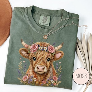 May include: Moss green Comfort Colors t-shirt featuring a detailed illustration of a cow with a floral crown. The cow has patchwork-style fur and is surrounded by flowers. A gold necklace rests on the shirt.
