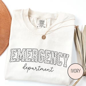 Puede incluir: Camiseta de color marfil con las palabras "EMERGENCY department" en negro. La palabra "EMERGENCY" está en una fuente punteada. Un collar dorado con un colgante está drapeado sobre la camiseta. La camiseta está doblada sobre una superficie blanca.