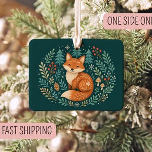 May include: A rectangular dark green ornament featuring a central illustration of a fox surrounded by a wreath of pine trees, berries, mushrooms, and snowflakes. The ornament hangs from a white ribbon and includes the text ONE SIDE ONLY.